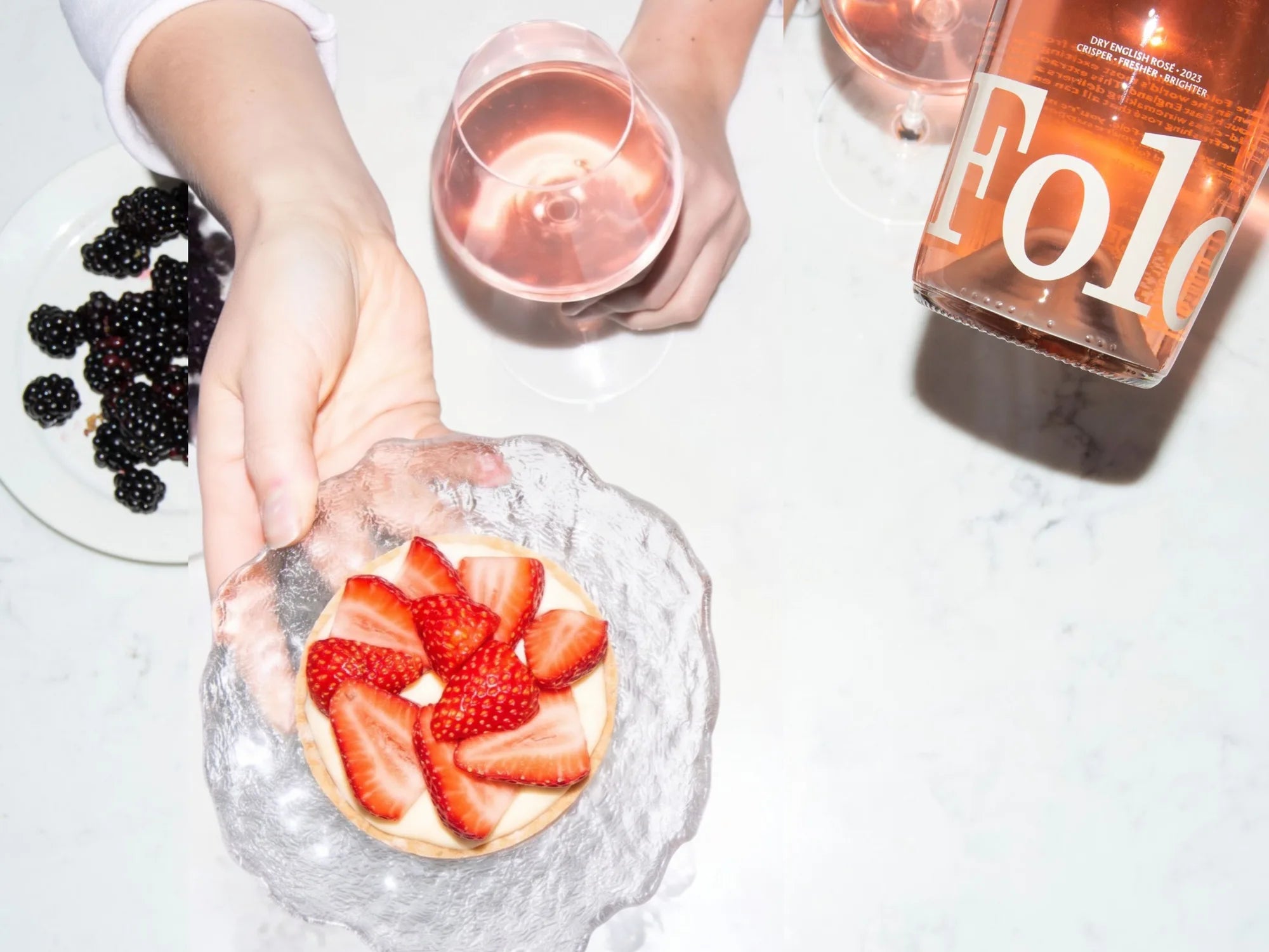 A hand holding a strawberry tart beside a glass and bottle of Folc rosé