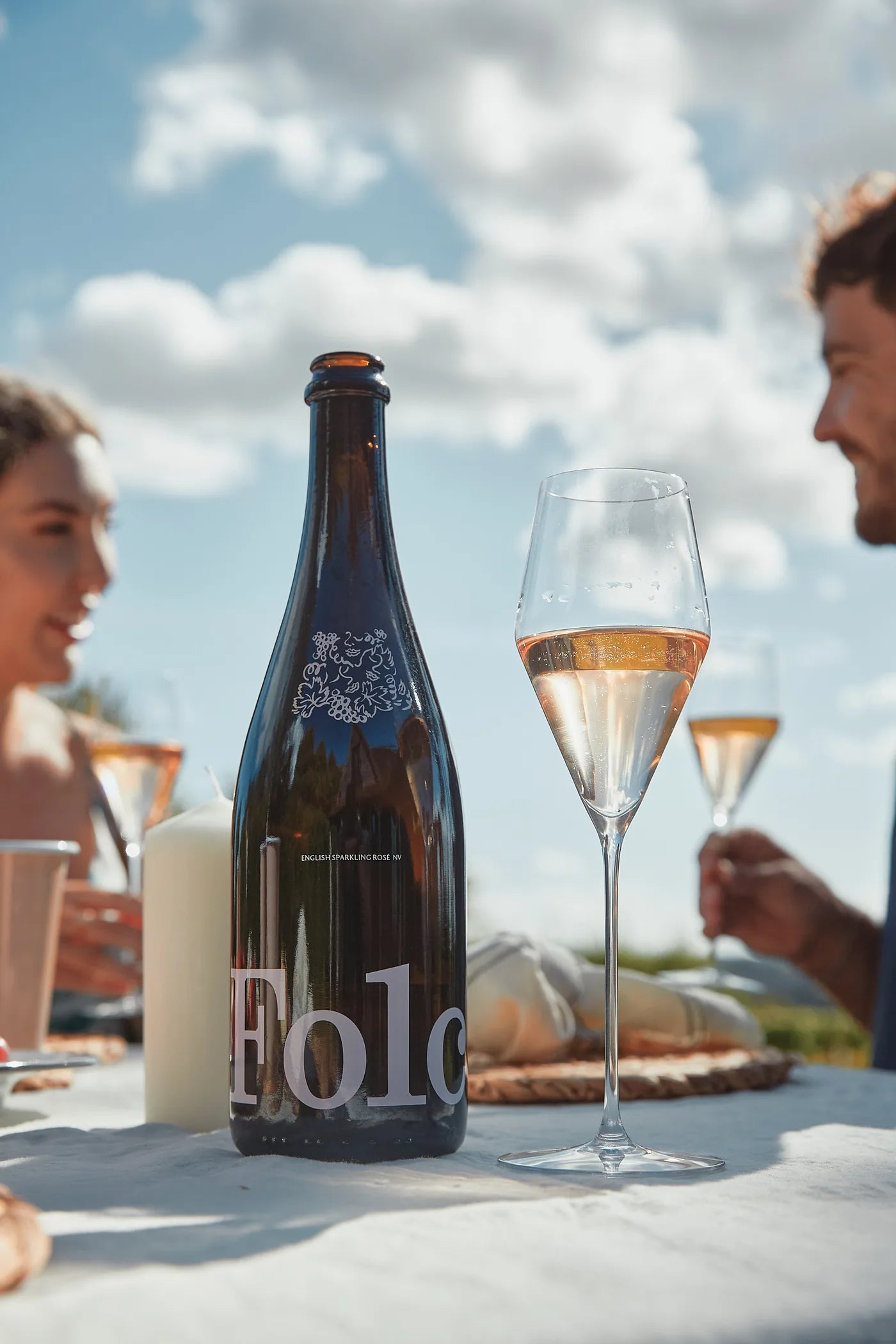 A bottle of Folc English Sparkling Rosé beside a flute of pale pink rosé on a picnic table