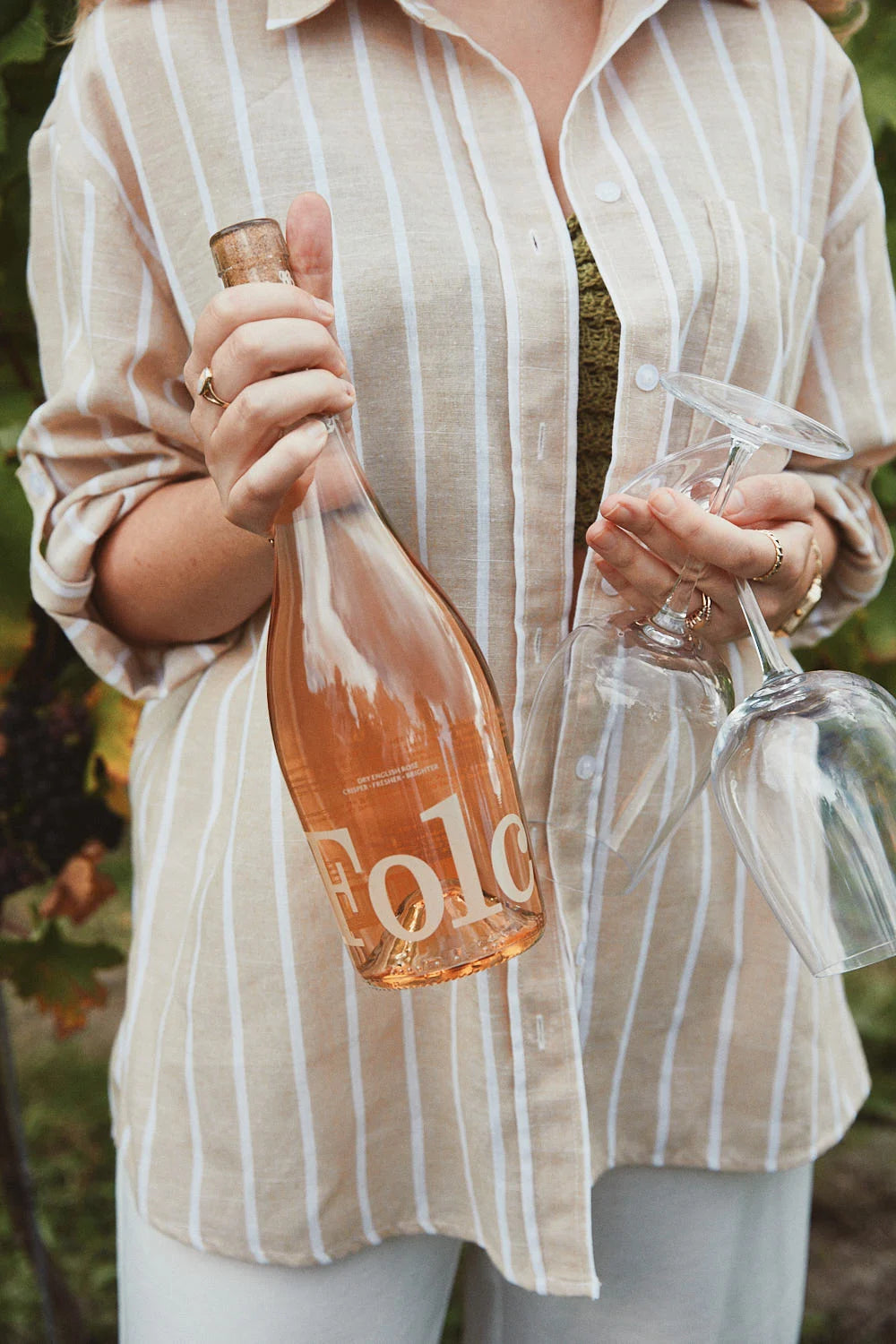 Someone in a striped shirt holding a bottle of Folc English Rosé and two glasses in a vineyard